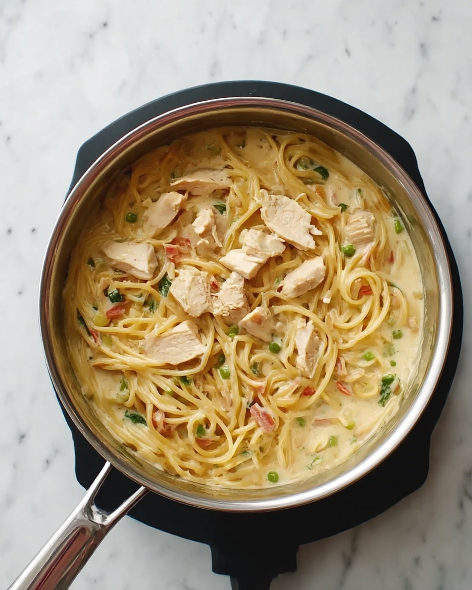 A silver pot filled with spaghetti in a creamy light-colored sauce with small pieces of green vegetables and bits of red scattered throughout. On top, there are several pieces of white chicken meat placed unevenly over the noodles. The pot is set on a black hot plate, all resting on a white marbled surface. photo taken with an iphone --ar 4:5 --v 7