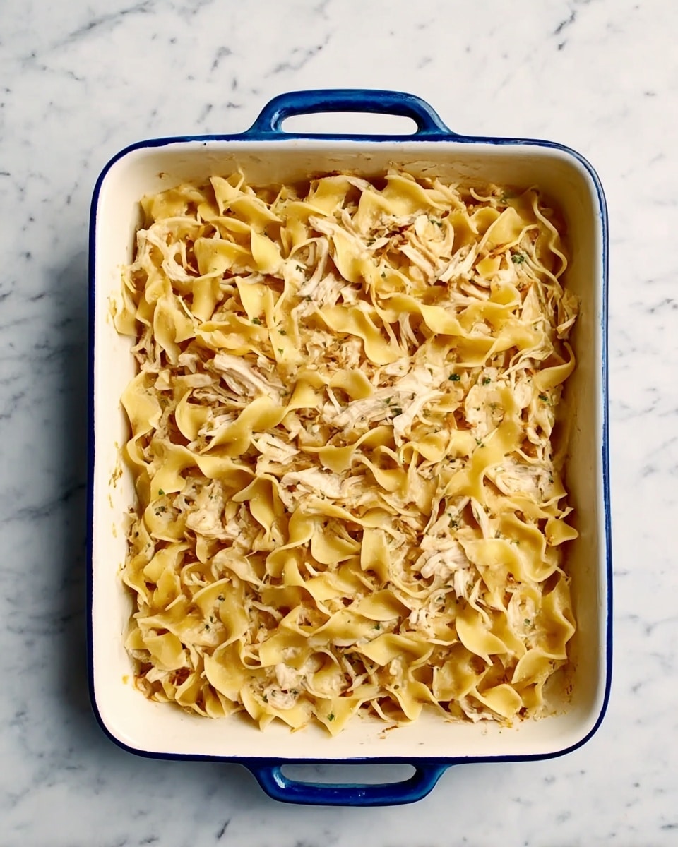 A white rectangular baking dish with blue handles holds a one-layer dish of wide egg noodles mixed with shredded chicken. The noodles are light golden yellow and soft-looking, scattered unevenly throughout. The shredded chicken is white with some darker bits, mixed gently within the noodles. The dish sits on a white marbled surface. photo taken with an iphone --ar 4:5 --v 7