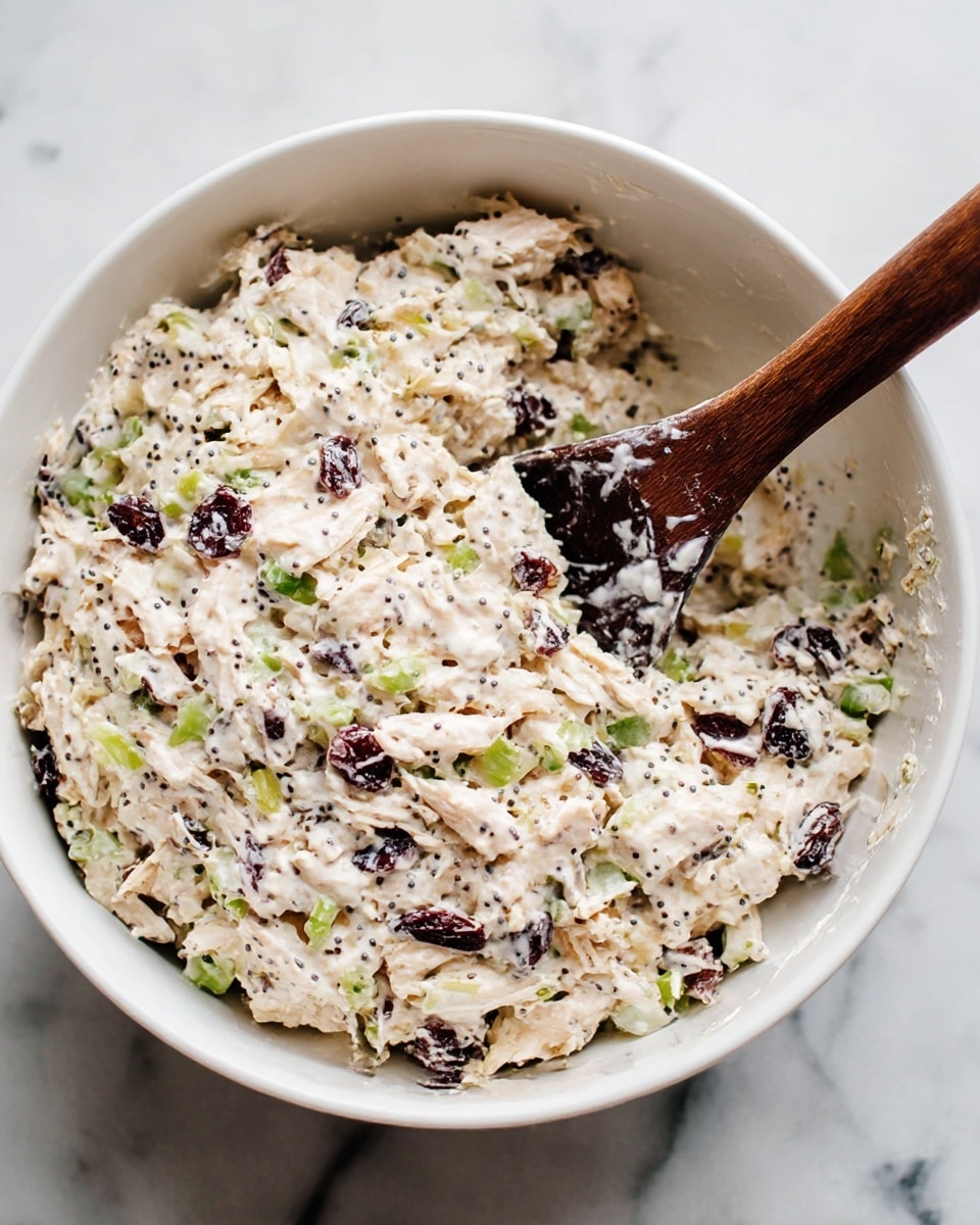 A white bowl filled with a creamy mixture that has visible pieces of light beige shredded chicken, dark dried cranberries, and bright green chopped celery, all mixed together with a white mayonnaise-like dressing that coats every ingredient. Small black poppy seeds are scattered throughout the dish, adding tiny specks of contrast. A dark wooden spoon rests inside the bowl, partially covered in the creamy mixture. The bowl is set on a white marbled surface. photo taken with an iphone --ar 4:5 --v 7