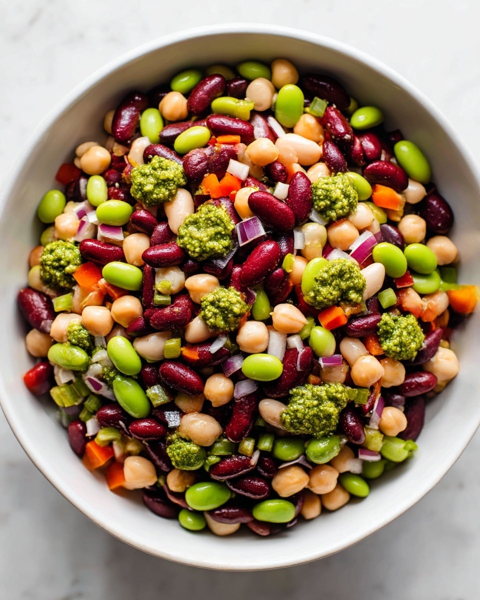 A close-up view of a colorful bean salad in a white bowl placed on a white marbled surface. The salad has many layers of mixed beans including dark red kidney beans, black beans, creamy white beans, and light brown chickpeas, all evenly spread throughout. These beans are mixed with diced small pieces of red onion, bright green edamame, orange bell pepper, and some bits of red bell pepper. On top, small dollops of green pesto sauce add a fresh, textured layer. The salad looks fresh with vibrant colors and a mix of smooth and slightly rough textures. Photo taken with an iphone --ar 4:5 --v 7