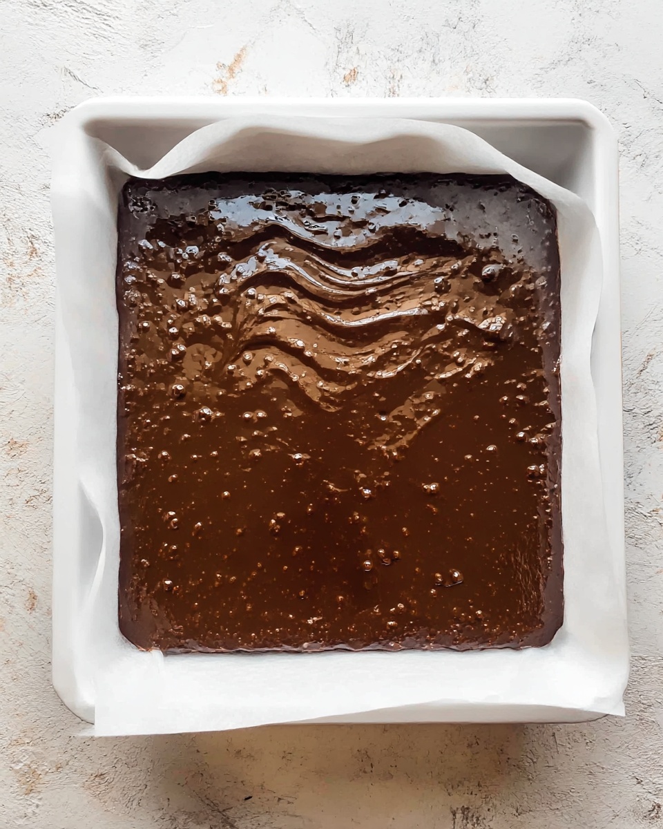A square white baking tray lined with white parchment paper holds a single layer of dark brown, shiny chocolate batter with a smooth but slightly bumpy texture on top, spread evenly to the edges. The surface shows small bubbles and a wavy pattern in the center where the chocolate may have been swirled or smoothed. The tray sits on a white marbled textured surface. photo taken with an iphone --ar 4:5 --v 7