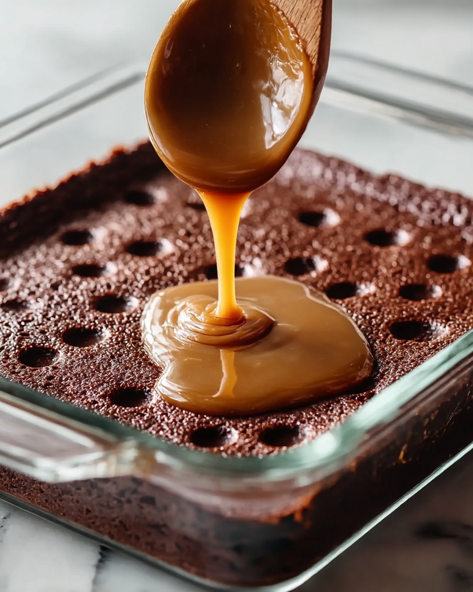 The image shows a close-up of a single thick, rich, dark brown chocolate cake layer in a clear glass square baking dish. The surface of the cake has many small round holes evenly spaced on top. A wooden spoon is held above, dripping a smooth, shiny light brown caramel sauce that is slowly falling onto the middle of the cake’s textured top, filling some of the holes with a glossy layer of caramel. The dish is set on a white marbled surface, and the scene is lit with soft natural light. Photo taken with an iphone --ar 4:5 --v 7