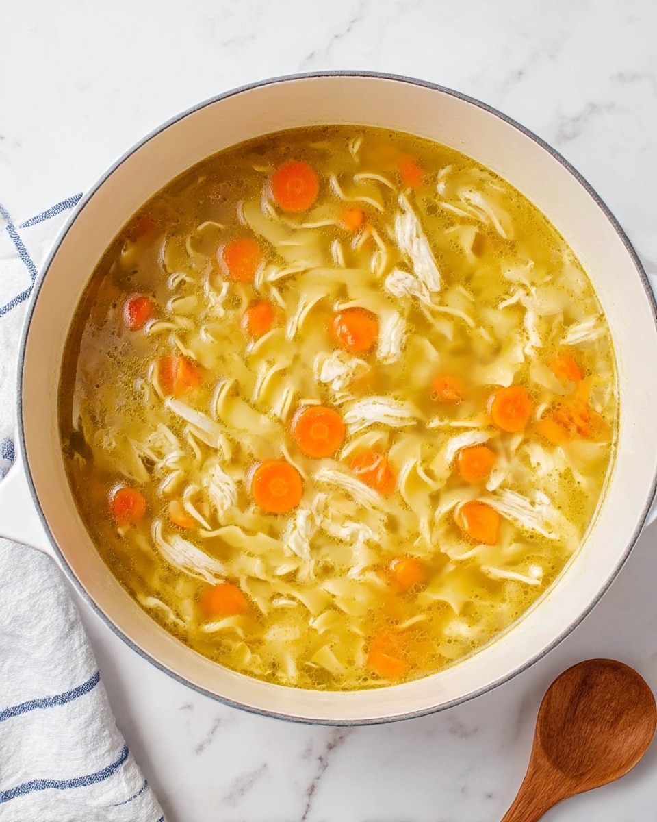 A white pot filled with clear golden broth soup containing three main layers: at the bottom, pale yellow egg noodles curled and loose; floating above the noodles, light orange carrot pieces cut into small chunks; scattered throughout the soup, white shredded chicken pieces. The soup has a glossy, smooth surface. The pot sits on a white marbled texture surface, with a white and blue striped cloth near the top left and a wooden spoon to the bottom right. Photo taken with an iphone --ar 4:5 --v 7