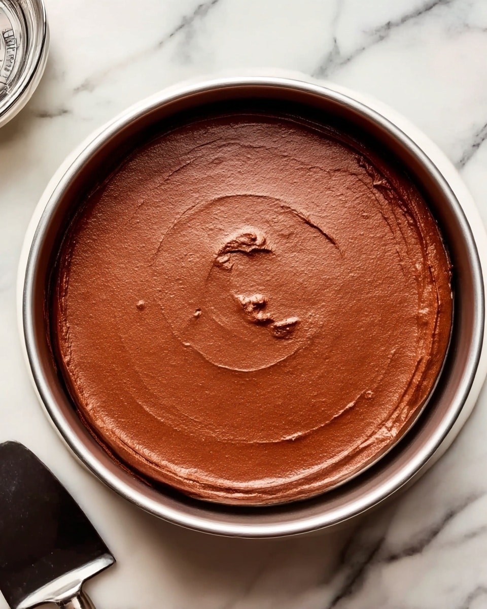 The image shows a round pan filled with smooth, thick chocolate batter evenly spread inside, creating one single dark brown layer. The pan is silver and shiny, resting on a pot with a dark kitchen tool next to it on a white marbled surface. The batter has a few small textures and swirls on the top, giving it a slightly glossy look. photo taken with an iphone --ar 4:5 --v 7