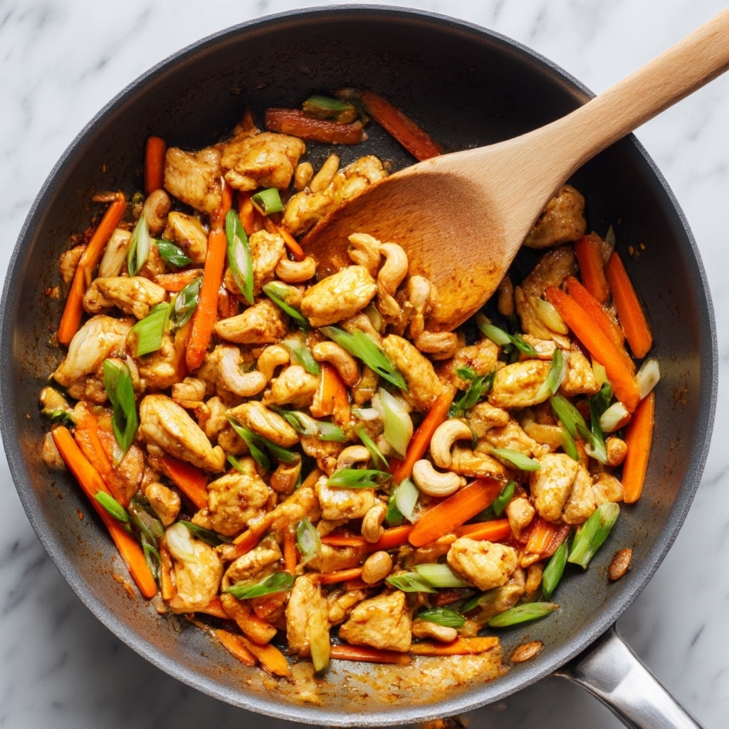 The image shows a pan filled with cooked pieces of golden brown chicken scattered evenly throughout. Thin, bright orange carrot sticks are mixed well among the chicken, adding a vibrant touch. Small green slices of scallions are spread across the dish, offering a fresh contrast in color. Whole cashew nuts intersperse the mixture, giving a crunchy texture appearance. A wooden spoon with light tan color is placed in the pan, partially covered by the food. The pan itself has a smooth, neutral gray non-stick surface. The background is white marble textured. photo taken with an iphone --ar 4:5 --v 7