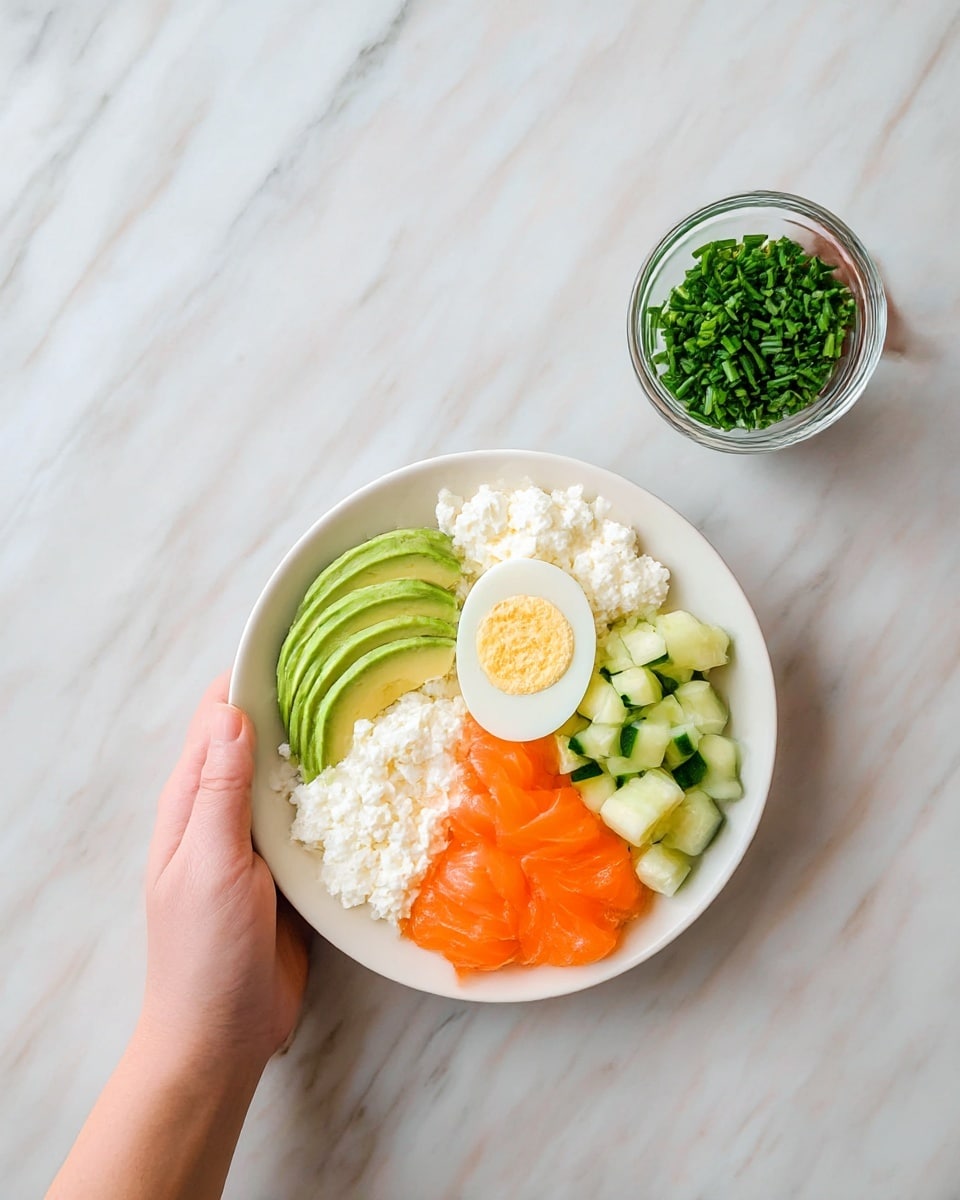 A white bowl holds five layers of food arranged side by side. Starting from the left, there is a thick layer of white cottage cheese with a soft, creamy texture. Next to it, slices of green avocado are fanned out smoothly. Beside the avocado, a half of a boiled egg shows a white outer part and a yellow yolk. To the right of the egg, orange slices of smoked salmon are loosely folded in delicate waves. Finally, small green cucumber cubes fill the right side of the bowl. A woman's hand is holding a small glass container filled with chopped green chives above the bowl. The bowl is set on a white marbled surface. photo taken with an iphone --ar 4:5 --v 7