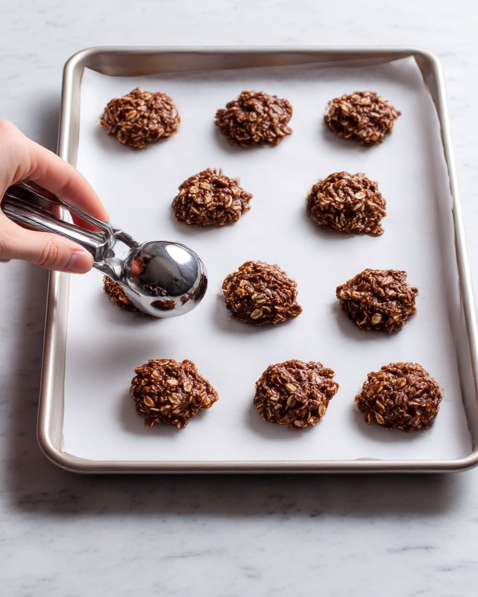 A baking tray lined with white paper holds nine round clusters of chocolate oatmeal cookie dough, arranged in three rows and three columns. The cookie dough has a rough texture and dark brown color with visible oats mixed throughout. A woman's hand holds a silver ice cream scoop, pressing down slightly on one of the dough clusters near the lower left corner of the tray. The tray sits on a white marbled surface. photo taken with an iphone --ar 4:5 --v 7