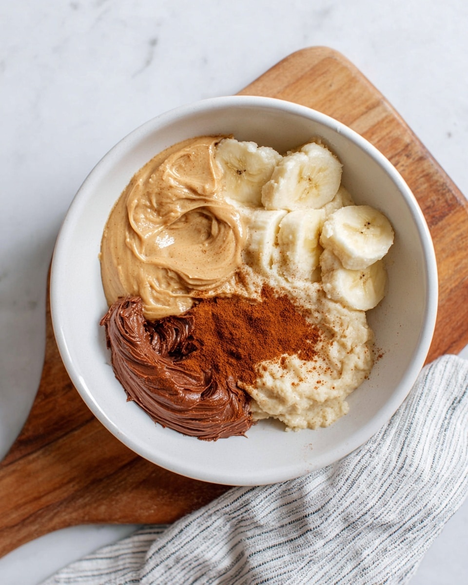 A white bowl on a wooden board with a striped cloth beside it contains four layers: mashed banana at the top right with a soft, creamy texture and pale yellow color, creamy peanut butter with a smooth texture and light brown color at the bottom right, soft chocolate spread with a rich brown color and smooth texture at the bottom left, and a sprinkle of cinnamon powder with a fine texture and reddish-brown color dusted near the chocolate spread. The bowl sits on a white marbled surface. Photo taken with an iphone --ar 4:5 --v 7
