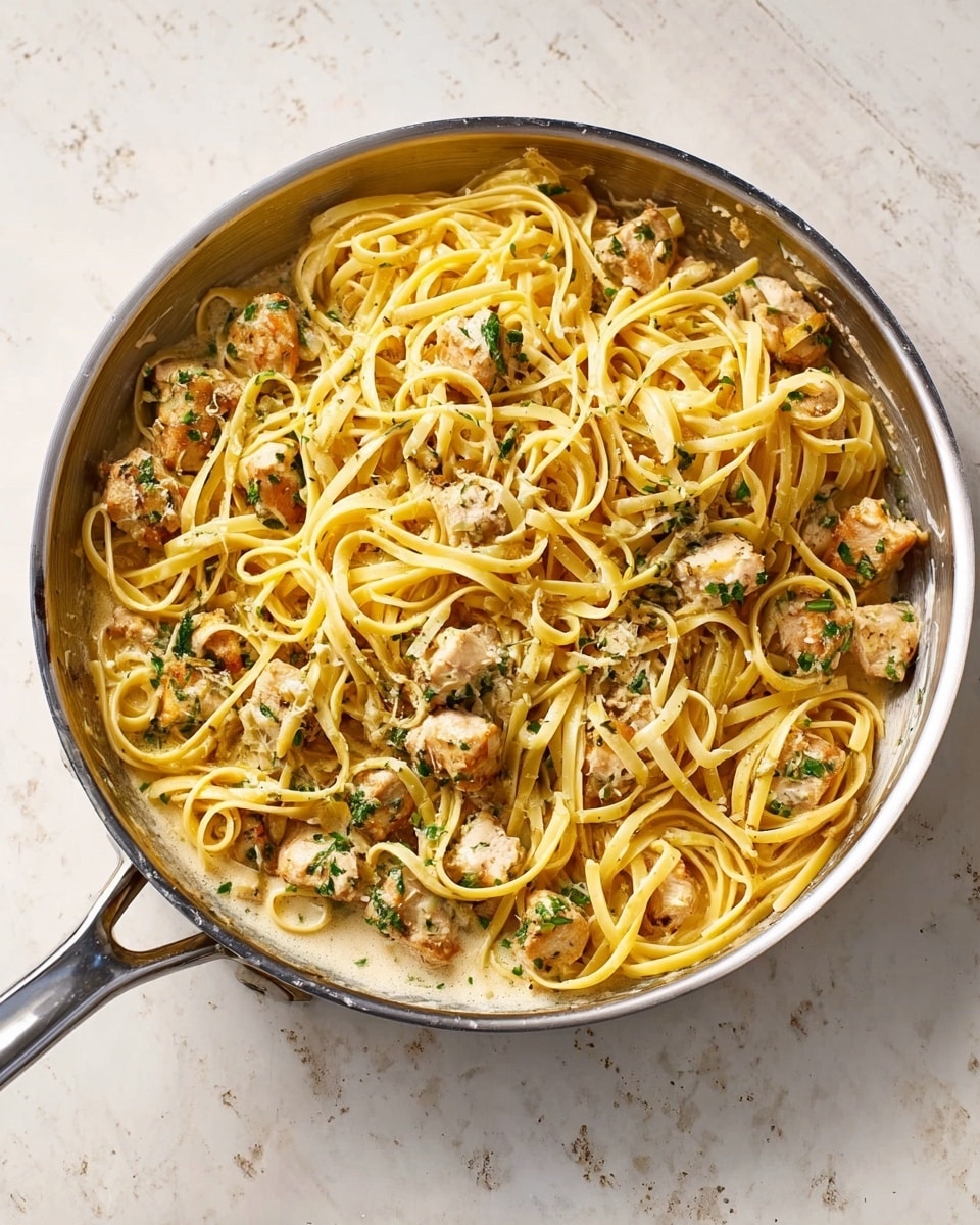 This image shows a silver pan filled with creamy chicken pasta. The dish has two main layers: a bottom layer of creamy light sauce, visible around the edges, and a top layer of thick, long yellow noodles mixed with chunks of cooked chicken that are light brown with green herbs sprinkled on them. The pan is placed on a white marbled surface. Photo taken with an iphone --ar 4:5 --v 7