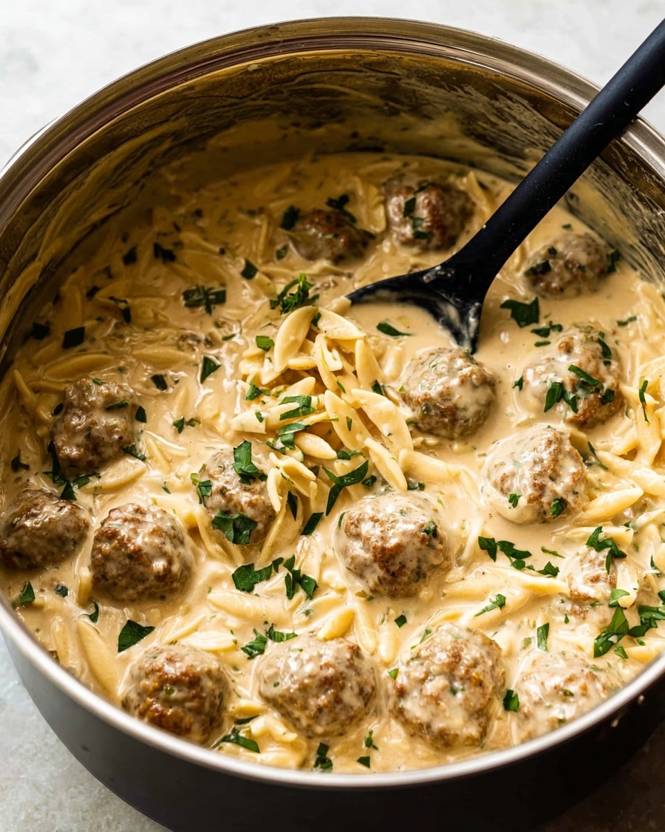 A close-up view of a large pot filled with creamy beige sauce mixed with small meatballs and short pasta pieces. The sauce looks smooth and thick, evenly coating the meatballs and pasta. Bright green herb leaves are sprinkled over the dish, adding a fresh contrast to the light sauce. A black spoon is partially submerged, ready for stirring. The pot's inner side shows traces of the sauce. The scene sits on a white marbled surface. photo taken with an iphone --ar 4:5 --v 7