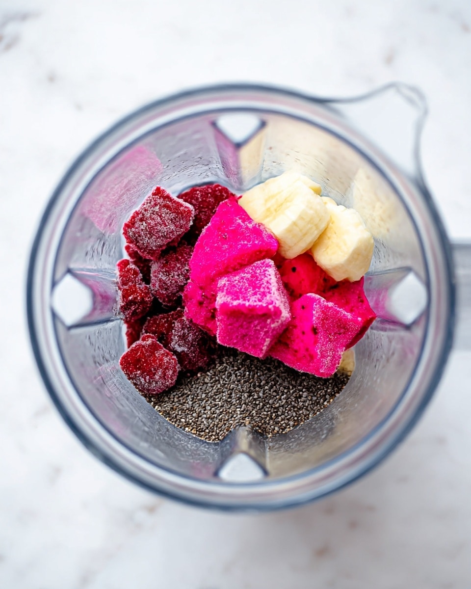 Inside a clear blender container, there are three layers of ingredients. The bottom layer consists of small black chia seeds scattered across the base. On top of this is a layer with frozen pieces of dark red strawberries, slightly frosty and textured. The top layer has bright pink frozen cubes of dragon fruit and pale yellow frozen banana chunks, both with a frosted texture, arranged together near the center. The blender sits on a white marbled surface. photo taken with an iphone --ar 4:5 --v 7