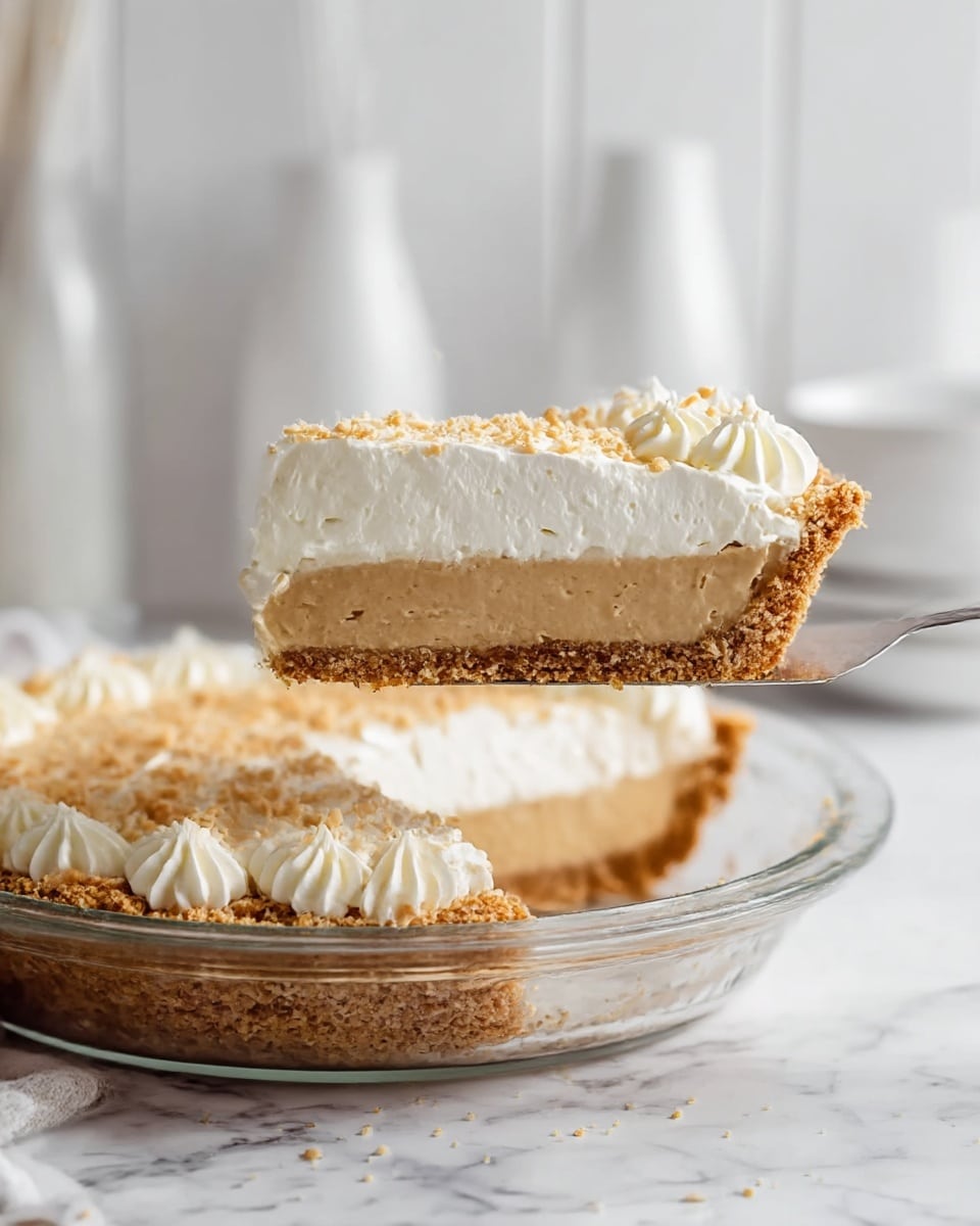 A slice of pie is lifted above a clear glass pie dish placed on a white marbled surface. The pie has three distinct layers: a crumbly golden brown crust forming the base and edges, a smooth light brown filling in the middle, and a thick white whipped topping with a slightly textured surface on top. The pie is decorated with small piped swirls around the edge under the whipped topping and sprinkled with fine crumbs, adding a slightly rough texture on the top. The background is softly blurred with white vases and bowls, enhancing the focus on the pie. Photo taken with an iphone --ar 4:5 --v 7