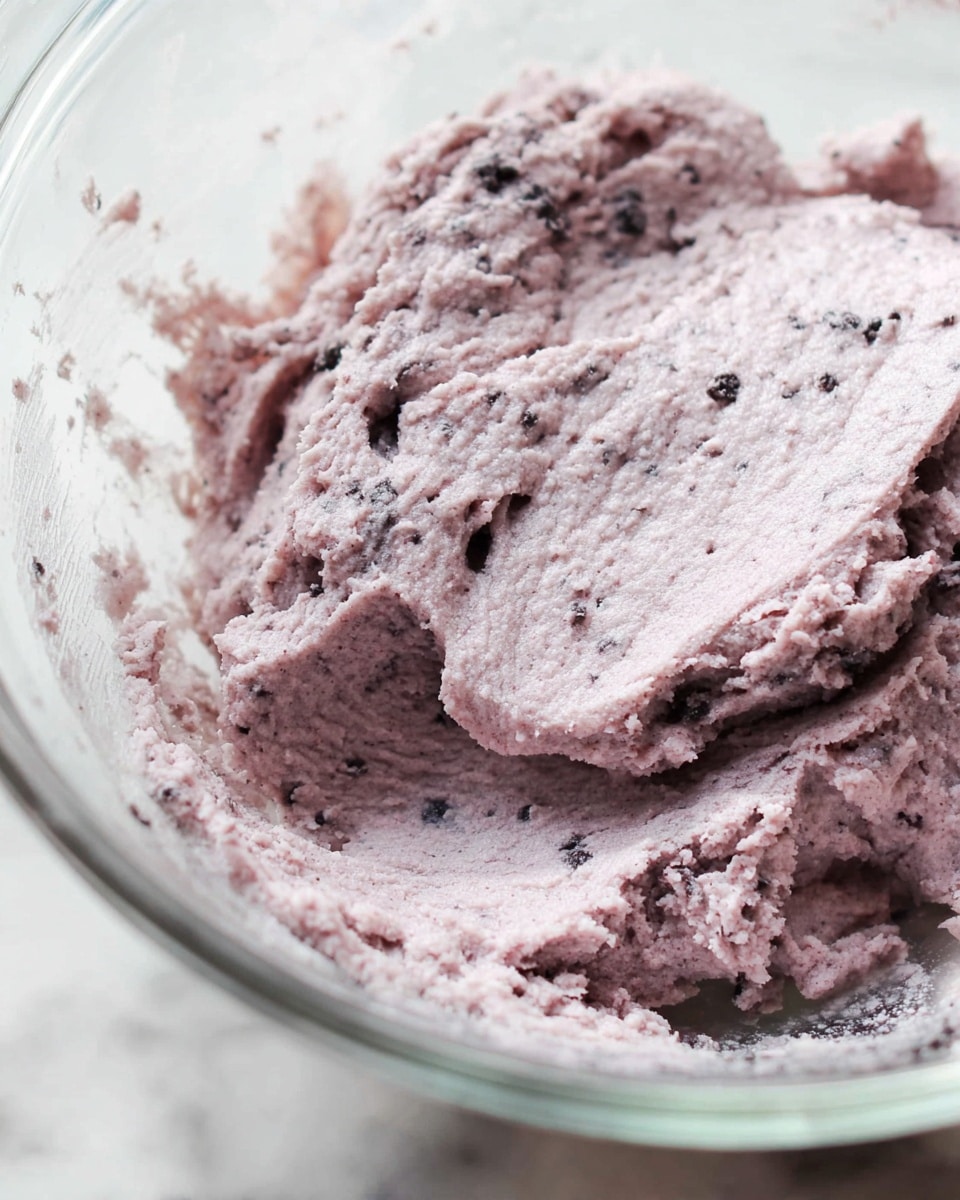 A close-up image shows a single layer of thick, light purple dough with small dark specks mixed throughout, spread unevenly inside a clear glass bowl. The dough has a rough, doughy texture with some lumps and soft peaks, sticking slightly to the sides of the bowl. The background is a white marbled surface, visible around the clear glass bowl. photo taken with an iphone --ar 4:5 --v 7