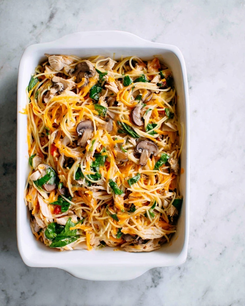 A white rectangular dish filled with a mixture of thin spaghetti noodles, shredded orange cheese, small pieces of cooked mushroom, shredded light brown chicken, and small green spinach leaves, all evenly layered together. The dish is placed on a white marbled surface, showing a top-down view with all ingredients mixed in a casual but colorful arrangement. photo taken with an iphone --ar 4:5 --v 7