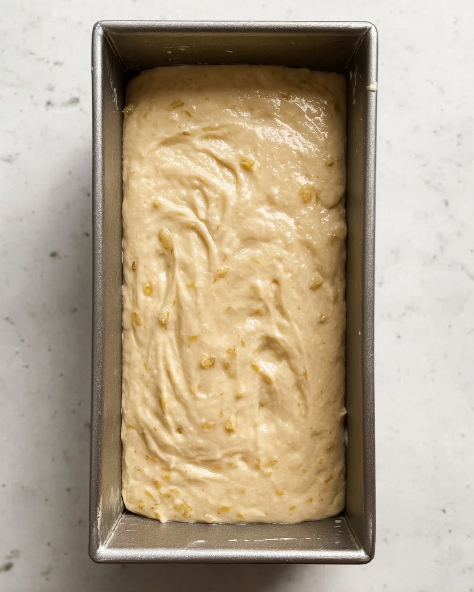 The image shows a metal rectangular baking pan filled with light beige batter that has a thick, smooth, and slightly lumpy texture. The batter spreads evenly across the pan, with some subtle swirled patterns on the surface, and tiny bits of lighter and darker beige visible throughout. The pan rests on a white marbled surface. photo taken with an iphone --ar 4:5 --v 7