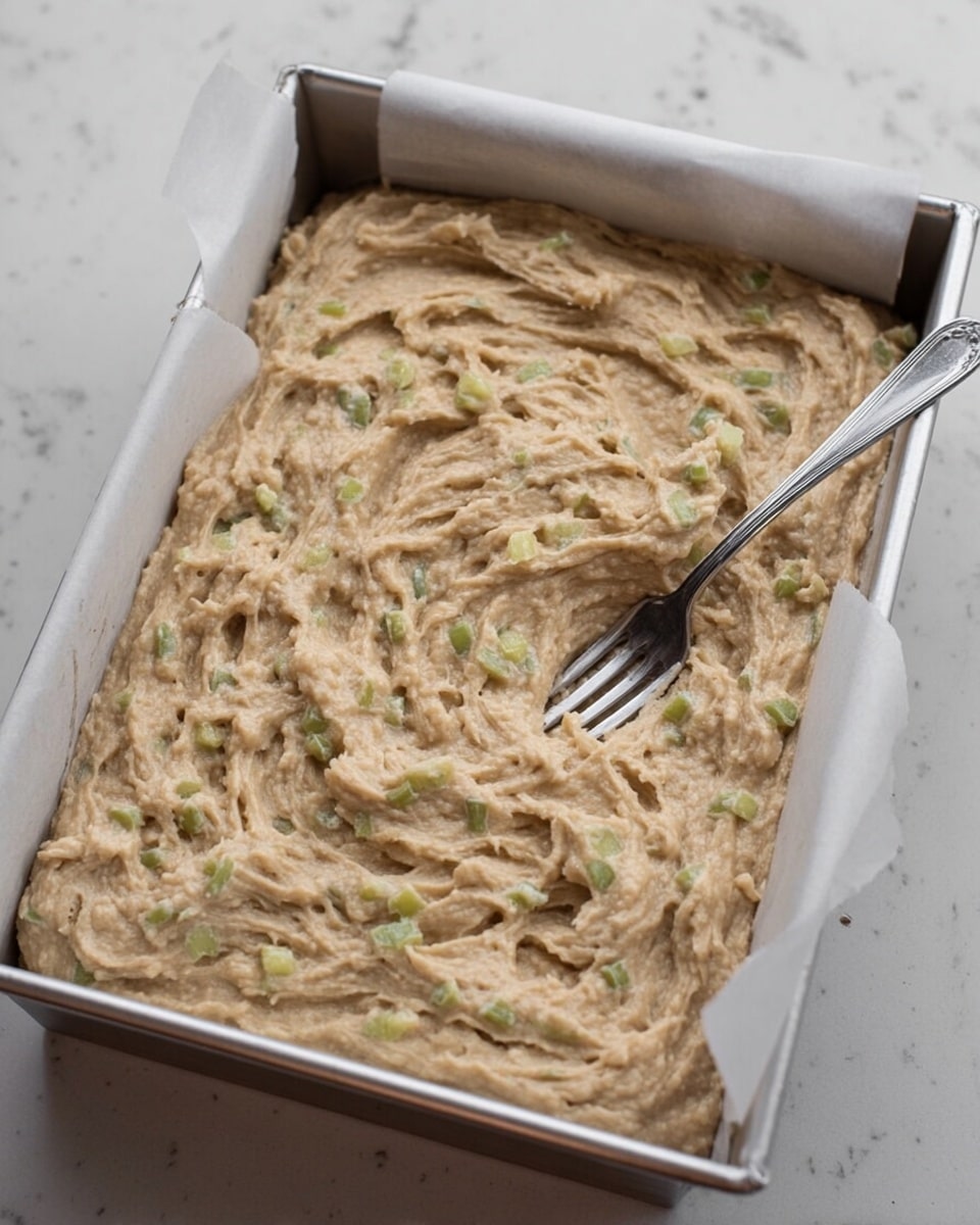 The image shows a silver rectangular baking pan on a white marbled surface, filled with a thick beige batter that has small greenish chunks mixed evenly throughout. The batter has a textured, swirled surface, created by a silver fork resting on top near the right side of the pan. The pan is lined with white parchment paper, which folds over the edges. The lighting is soft and natural, highlighting the creamy yet slightly chunky texture of the batter photo taken with an iphone --ar 4:5 --v 7