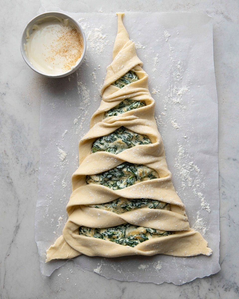The image shows an unbaked dough shaped like a Christmas tree with 9 horizontal layers of twisting branches on both sides filled with a green and white creamy spinach and cheese mix; the dough is light beige, slightly dusted with flour, and laid flat on parchment paper over a white marbled surface. Next to the dough tree, on the top left, is a small white bowl filled with a white creamy sauce topped with a light brown powder. The dough layers are folded outward in a twist style, revealing the filling clearly. photo taken with an iphone --ar 4:5 --v 7
