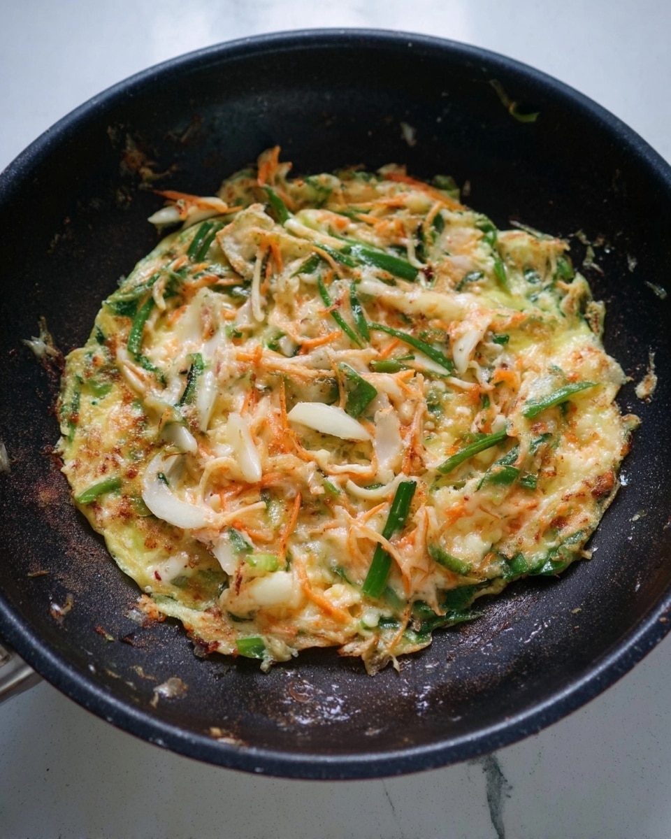 The image shows a single layer of mixed vegetable pancake cooking in a black pan. The pancake has green strips of vegetables, white curved pieces, and thin orange strands spread evenly. The texture looks soft with some parts starting to cook and stick together. The background and surface below the pan are a white marbled texture. photo taken with an iphone --ar 4:5 --v 7