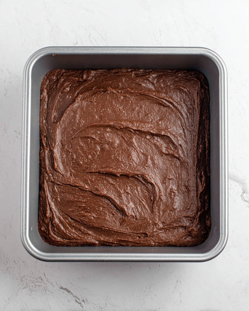 The image shows a square metal baking pan filled with smooth, thick chocolate batter. The batter has a dark brown color with slight uneven swirls and a soft texture on top, filling the entire pan evenly. The pan sits on a white marbled surface, and there is no other decoration or items around the pan photo taken with an iphone --ar 4:5 --v 7