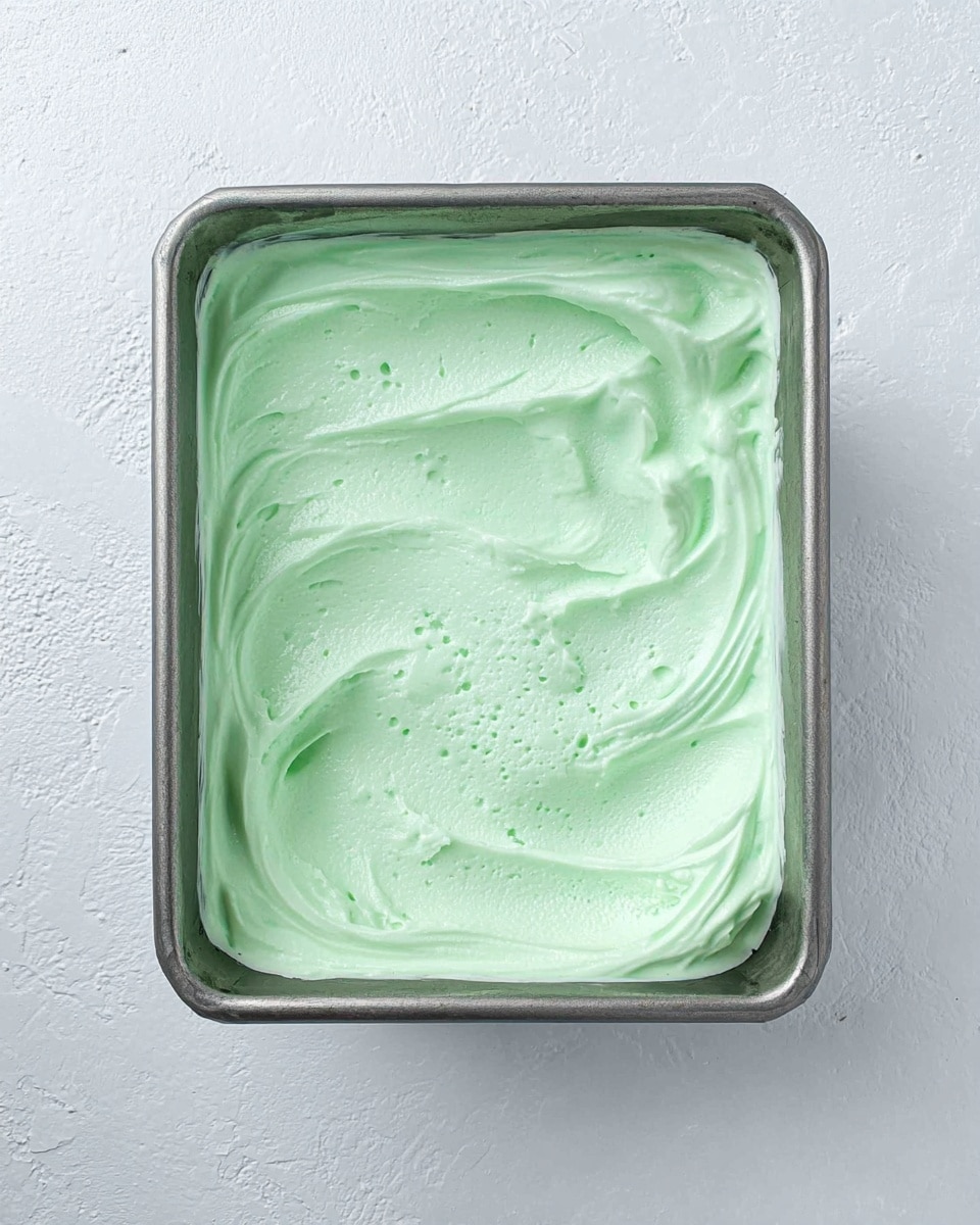 A square metal container filled with one thick layer of smooth, creamy light green ice cream with soft swirls and small air bubbles visible on the surface. The container sits on a white marbled textured surface. Photo taken with an iphone --ar 4:5 --v 7