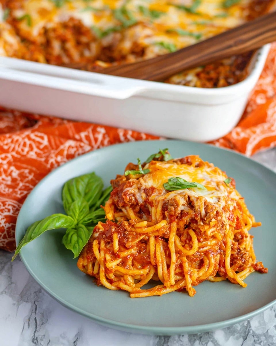 A serving of baked spaghetti is placed on a white plate, showing a layered mix of cooked spaghetti noodles covered in a rich red tomato sauce mixed with browned meat and melted golden cheese. A fresh green basil leaf rests on top as a garnish. In the background, a white casserole dish filled with the same spaghetti mixture topped with cheese is partly visible, sitting on an orange patterned cloth. The scene rests on a white marbled texture with a wooden spoon handle slightly visible. Photo taken with an iphone --ar 4:5 --v 7
