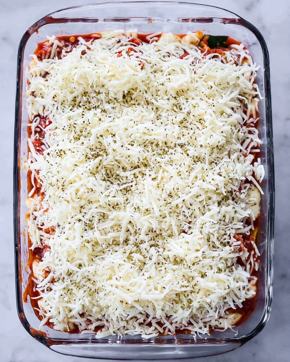 A clear glass baking dish filled with layered lasagna is shown from above. The top layer is a mix of shredded white cheese scattered evenly, with some greenish herb spices sprinkled throughout. Below the cheese, red tomato sauce peeks through in small areas, contrasting with the white cheese. The edges show hints of a green vegetable or herb layer beneath the cheese and sauce. The dish sits on a white marbled surface. photo taken with an iphone --ar 4:5 --v 7