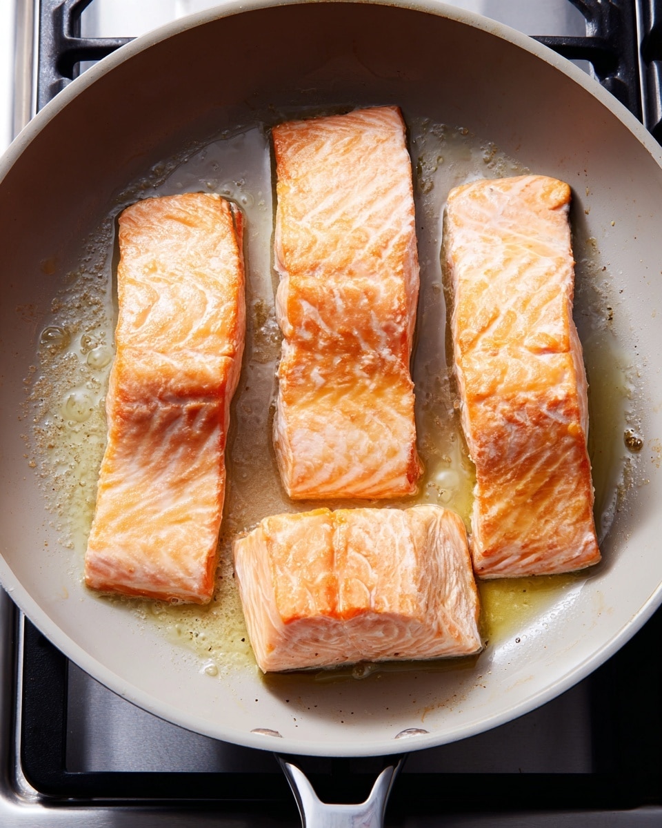 Four pieces of salmon fillets are cooking in a large light gray pan. The salmon pieces are arranged close but not touching; three pieces are aligned in a row at the top, and one longer piece is at the bottom. Each salmon piece has a light golden brown cooked layer on top, showing a soft and slightly shiny texture. The pan has some light oil around the salmon, making the surface slightly glossy. The background shows parts of a silver stove with black heating elements. Photo taken with an iphone --ar 4:5 --v 7