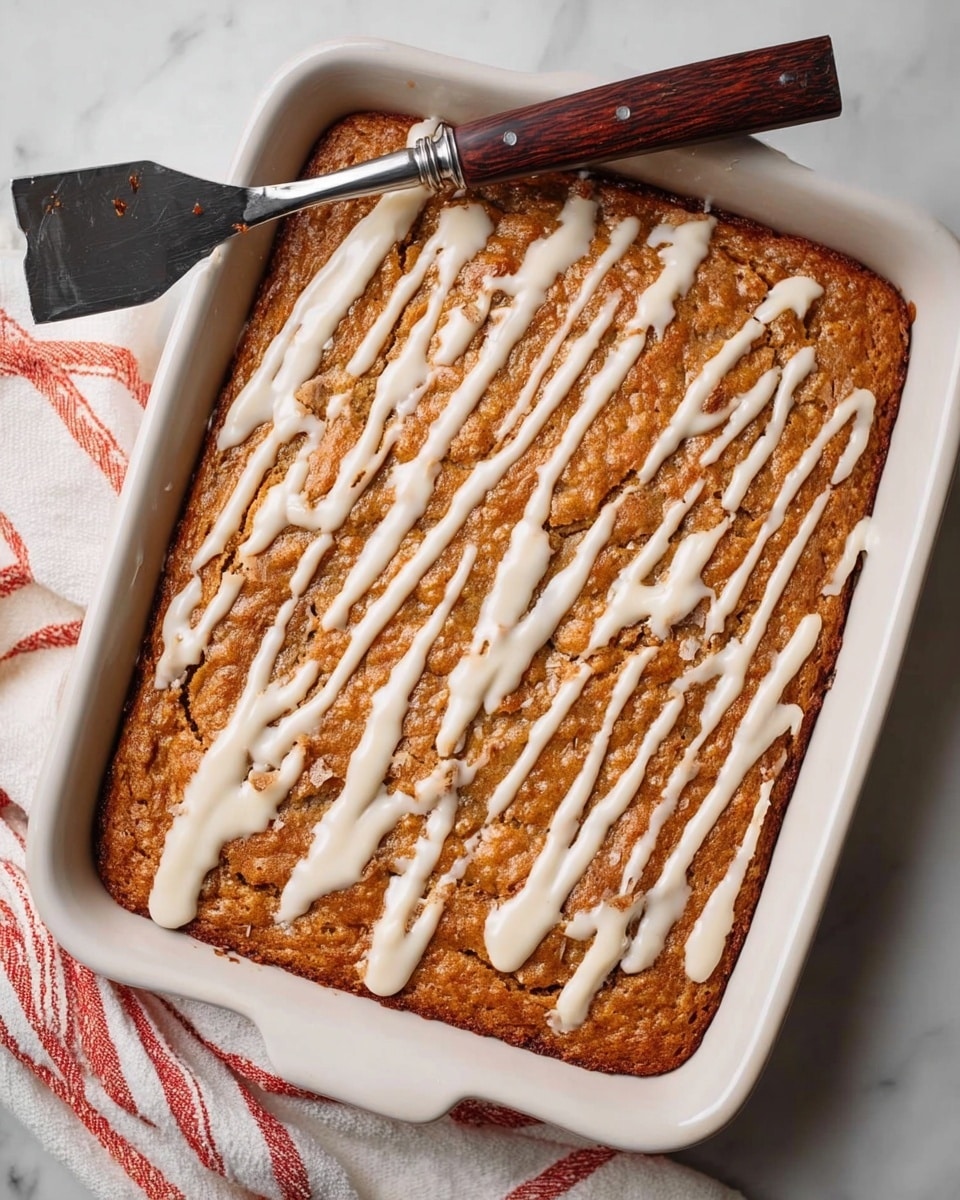 A rectangular baked dish with one thick layer of golden brown cake that has a slightly rough texture on top. It is drizzled with uneven white icing in thin and thick lines across the entire surface. The dish is white and sits on a white marbled surface. A metal spatula with a dark brown wooden handle rests on the top left corner of the dish. A white cloth with red stripes is partially visible under the bottom left corner of the dish. photo taken with an iphone --ar 4:5 --v 7