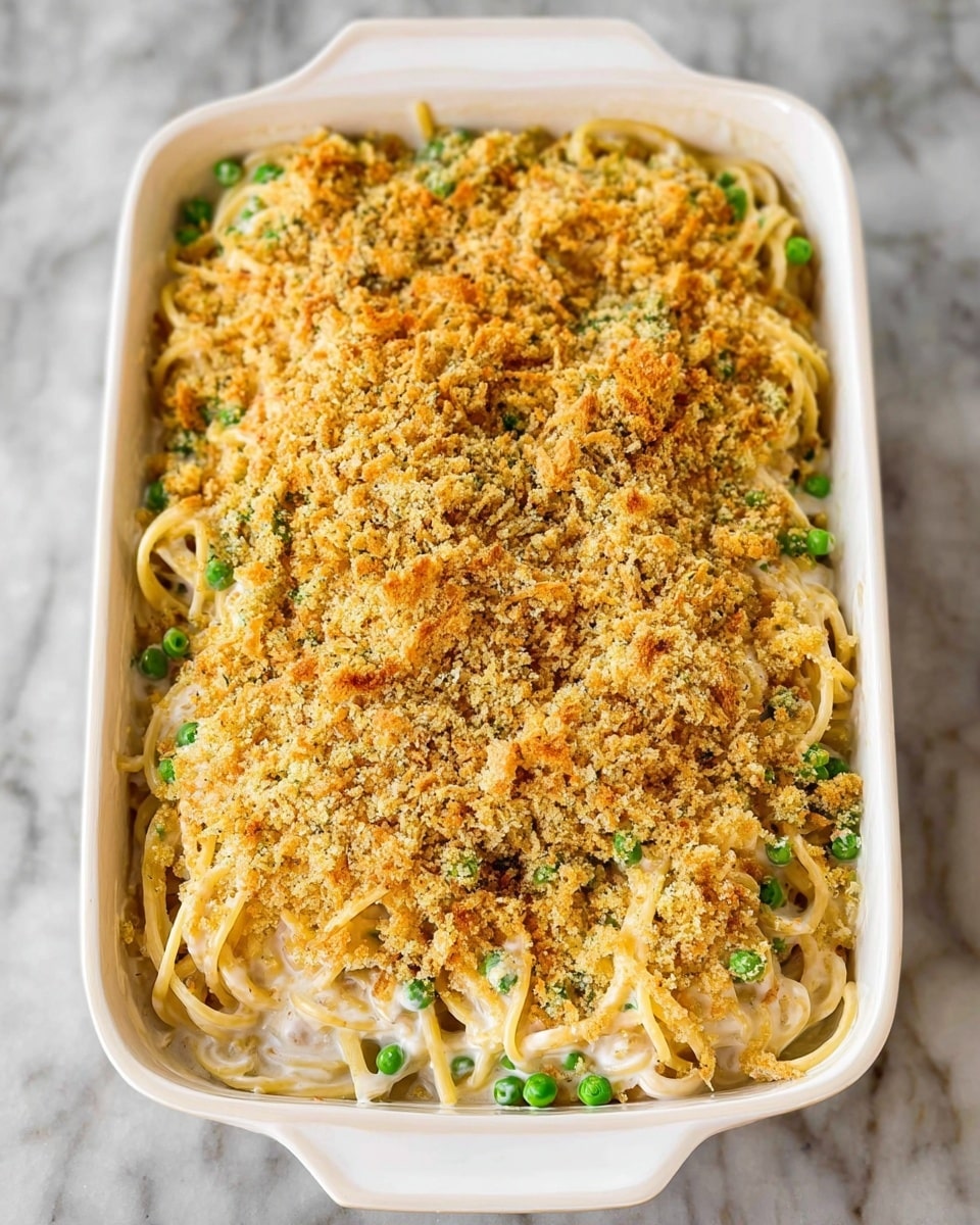 A white rectangular dish filled with a layered pasta casserole placed on a white marbled surface; the bottom layer consists of spaghetti noodles mixed with creamy white sauce and green peas, creating a soft and chunky texture. On top of this is a thick layer of golden brown, crunchy breadcrumb topping, spread evenly to cover the pasta completely, with some strands of noodles peeking through the edges. The casserole looks warm and inviting, with a mix of creamy and crispy textures visible. photo taken with an iphone --ar 4:5 --v 7