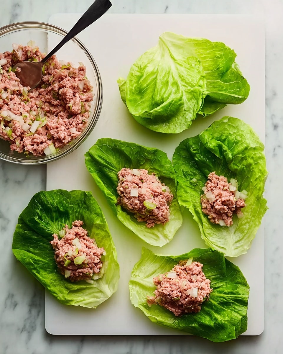 The image shows a white cutting board on a white marbled surface. On the cutting board, there are five large, fresh green lettuce leaves arranged in two rows. Three leaves in the front row each hold a mound of raw pinkish ground meat mixed with small white pieces, likely onions. Two empty lettuce leaves are placed in the back row. To the top left of the cutting board, a clear glass bowl contains more of the same meat mixture with a black-handled metal spoon resting inside. The scene is bright and clean, with fresh ingredients ready to be used. Photo taken with an iphone --ar 4:5 --v 7