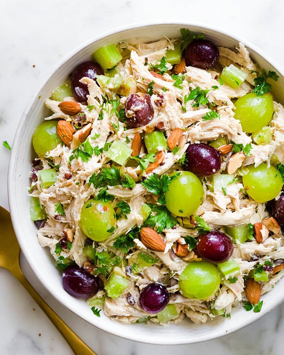 A large white bowl filled with shredded light beige chicken layered with bright green celery slices, whole and halved dark purple grapes, and small chunks of chopped light brown almonds. Fresh, finely chopped vibrant green parsley is scattered generously on top, adding a lively touch. The bowl is placed on a white marbled surface, with a few almond pieces nearby, and a gold spoon slightly visible on the left side. The overall texture is a mix of creamy chicken, crunchy celery and almonds, and juicy grapes, making for a colorful and fresh salad photo taken with an iphone --ar 4:5 --v 7