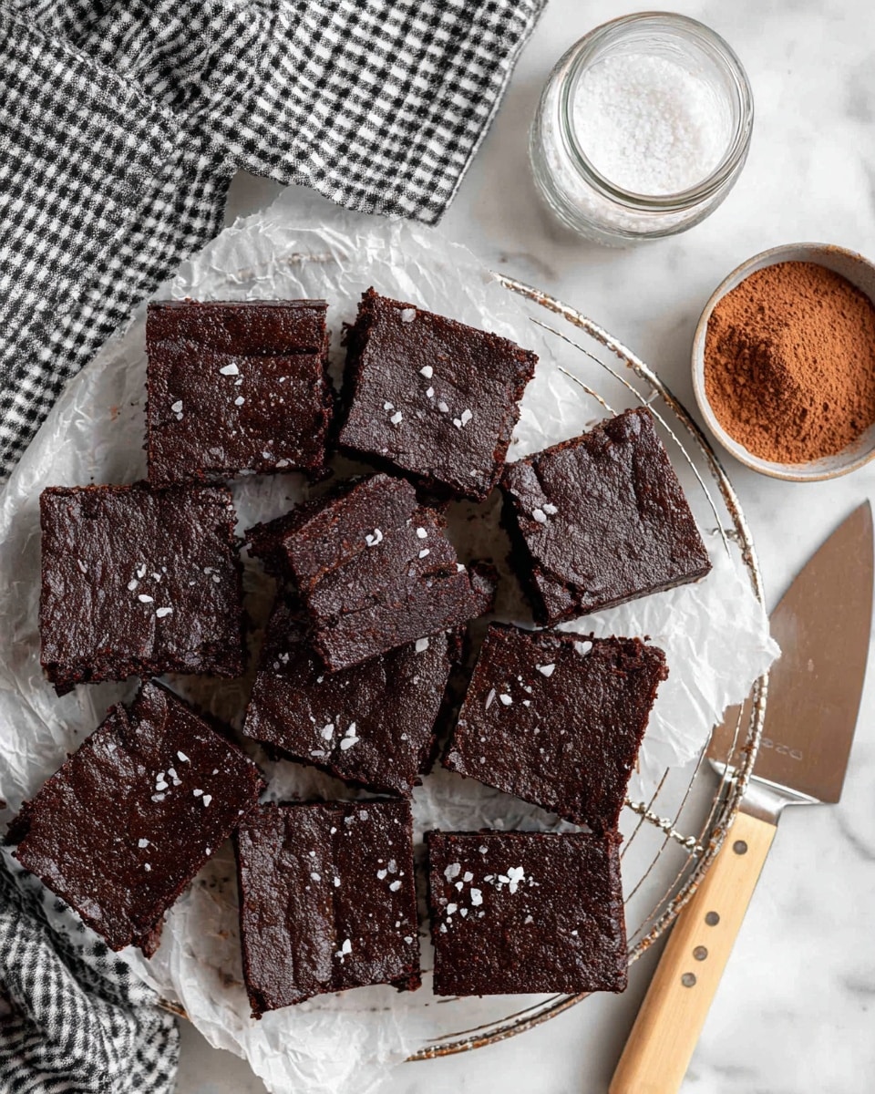 The image shows a batch of dark brown brownies cut into square pieces on a white parchment paper lined on a round wire cooling rack. Each brownie has a shiny, slightly crinkled top with small flakes of coarse white salt sprinkled over them. The edges appear a bit thicker and raised compared to the middle parts. Near the brownies are a small bowl of coarse salt and a glass jar with cocoa powder, both placed on a white marbled surface. A large spatula with a light wooden handle rests beside the cooling rack. At the top of the image, a black and white checkered cloth is partly visible. photo taken with an iphone --ar 4:5 --v 7