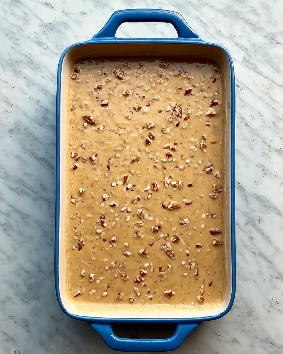 A rectangular blue-handled baking dish filled with a smooth, thick mixture of light brown color with small bits of nuts evenly spread throughout. The surface looks glossy and slightly textured with tiny nut pieces visible all over. The dish sits on a white marbled textured surface. photo taken with an iphone --ar 4:5 --v 7