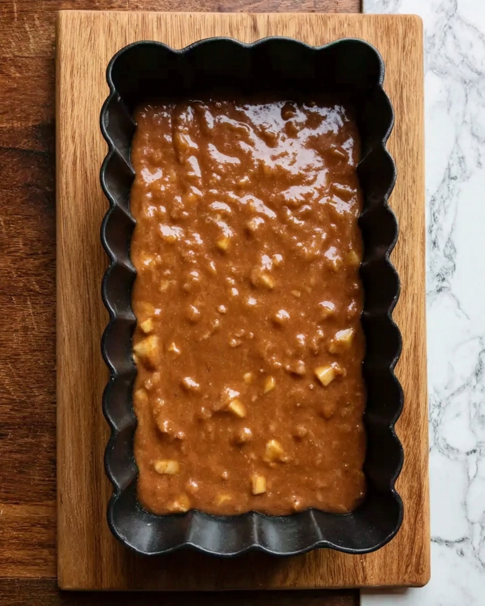 A black rectangular baking pan with scalloped edges is filled with a thick, brown batter mixed with small chunks of what looks like apple pieces spread evenly throughout. The pan sits on a wooden surface, but the background has been changed to a white marbled texture for the description purpose. The mixture looks smooth with some uneven texture from the fruit pieces inside. Photo taken with an iphone --ar 4:5 --v 7