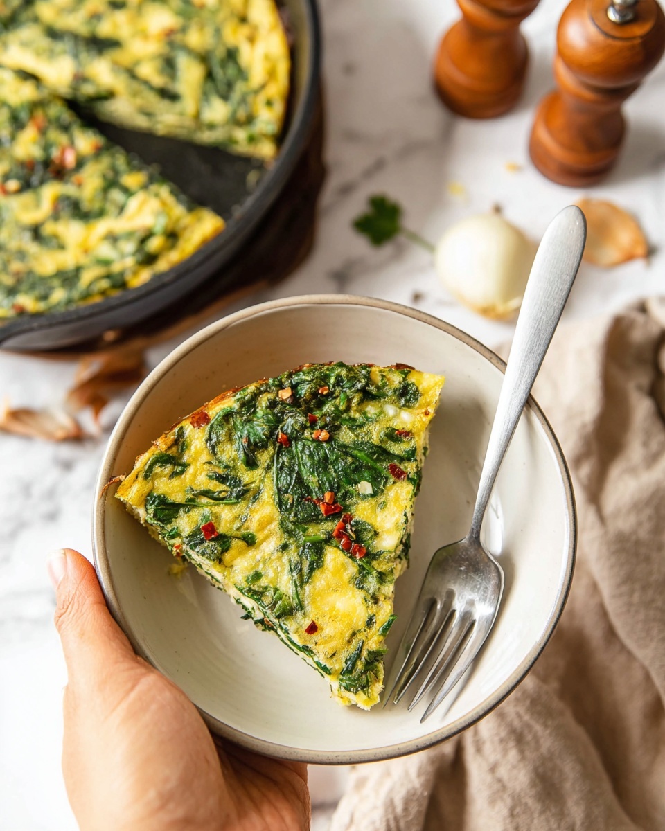 Crustless Spinach Quiche (Gluten-Free, Dairy-Free) Recipe 5 A slice of green spinach and yellow egg frittata fills most of a white round bowl, its surface showing visible layers of cooked spinach leaves mixed in a soft, slightly bumpy yellow egg base with small red chili flakes sprinkled on top. The slice is triangular, placed on the bottom left inside the bowl with a silver fork resting on the right side of the slice. A woman's hand holds the bowl from the left side. In the blurred background, there is a black skillet with the remaining frittata, a beige cloth, wooden pepper grinders, and onions on a white marbled surface. Photo taken with an iphone --ar 4:5 --v 7