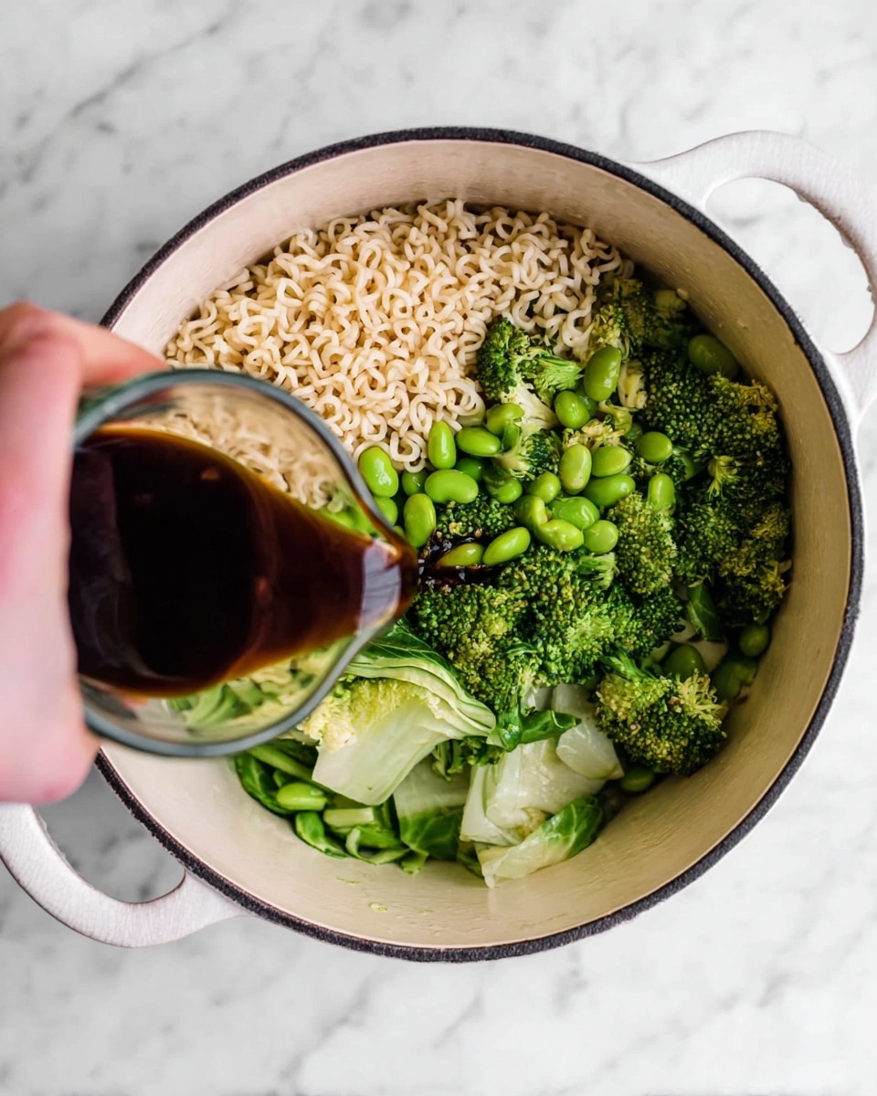 Inside a white pot with a black rim sits a mix of three layers: at the bottom, curly pale beige ramen noodles; on top of the noodles on one side, bright green broccoli florets and bok choy pieces with a leafy texture; scattered among them are small, round green edamame beans. A woman's hand is pouring a dark brown sauce from a clear glass cup onto the ingredients. The pot is placed on a white marbled surface. photo taken with an iphone --ar 4:5 --v 7