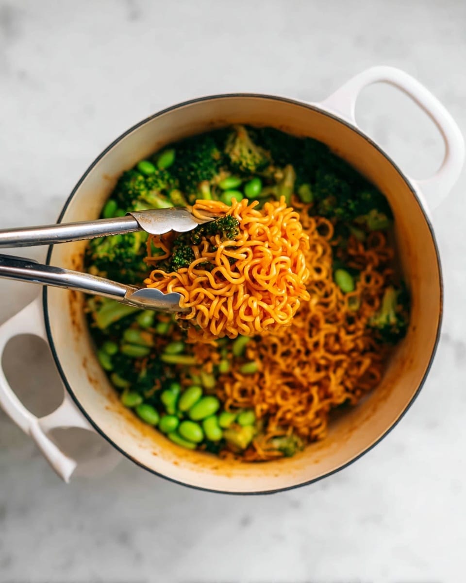 A white pot filled with cooked curly orange noodles placed on top, surrounded by bright green vegetables including broccoli and edamame beans. A pair of silver tongs holds some noodles and a piece of green vegetable above the pot. The pot sits on a white marbled surface, with a soft light highlighting the textures of the food and the pot. photo taken with an iphone --ar 4:5 --v 7