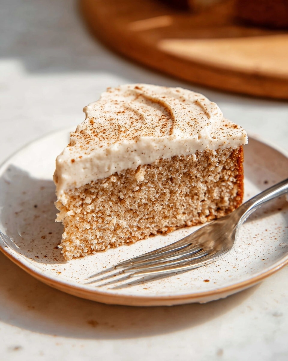 A single square slice of light brown cake with a soft, slightly crumbly texture sits on a round white plate. The cake has one visible layer topped with a thick, creamy frosting that is off-white with tiny brown specks, spread smoothly but with soft peaks. A light sprinkle of cinnamon or similar brown powder dusts the top of the frosting evenly. Next to the cake slice on the plate is a silver fork resting flat. The scene is set on a white marbled surface with warm, natural lighting highlighting the cake’s texture and the smoothness of the frosting. photo taken with an iphone --ar 4:5 --v 7