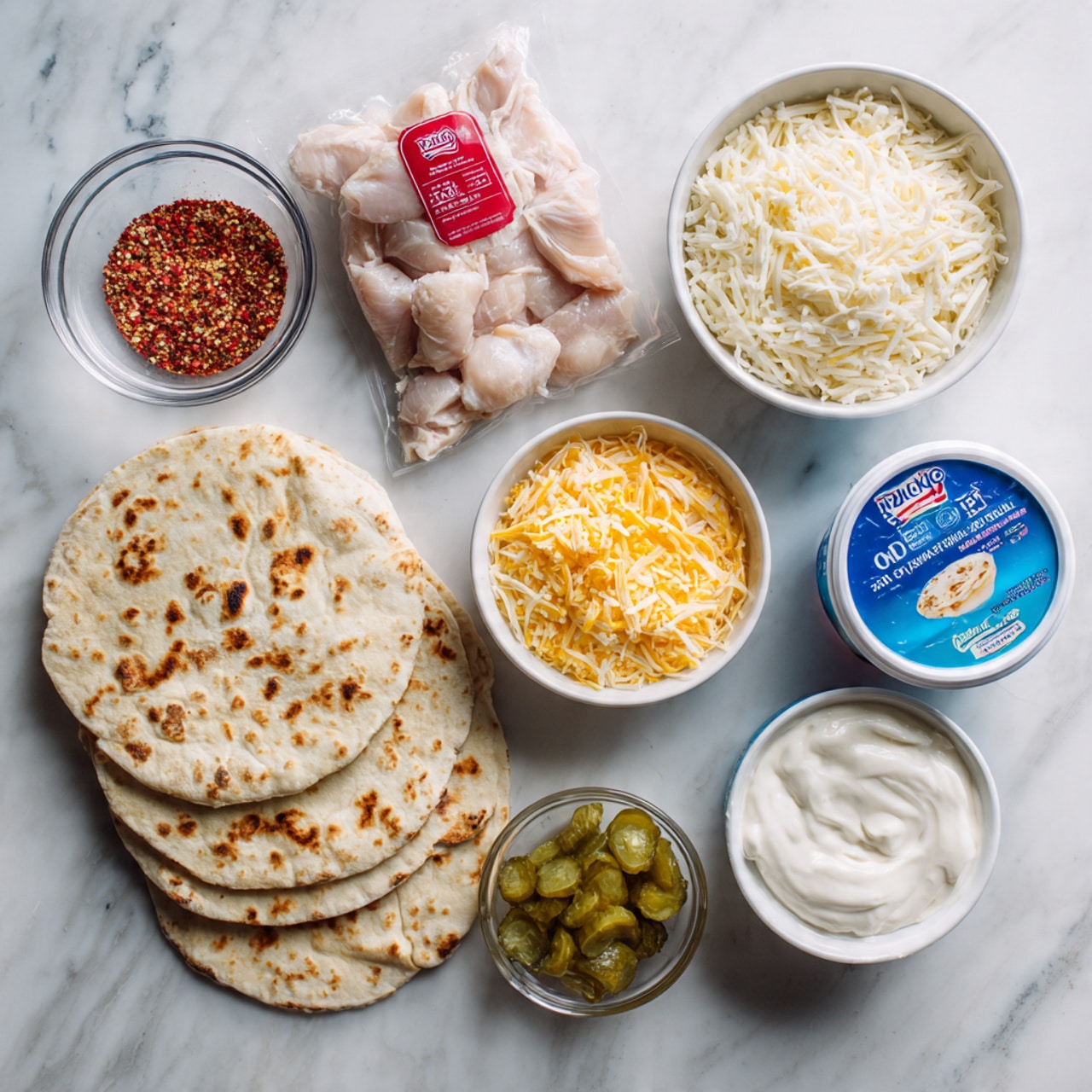 The image shows several ingredients arranged neatly on a white marbled surface. At the center bottom, in a stack, are five round flour tortillas with light brown spots. To the left of the tortillas, there is a small clear glass bowl filled with red and white seasoning mix. Above this bowl, there is a white bowl filled with shredded white cheese. At the top center, there is a clear plastic package containing raw, pale pink chicken pieces with a red label on top. To the right of the chicken, a white bowl holds shredded yellow cheese. Below the yellow cheese bowl, there is a blue and white container of Oikos 0% fat Greek yogurt, and beside that, a white tub of light sour cream with blue labeling. At the bottom right, a small clear glass bowl contains green chopped pickles in liquid. The photo taken with an iphone --ar 4:5 --v 7