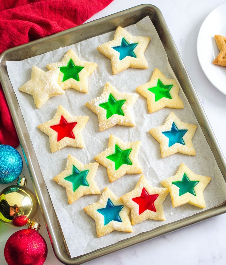 The image shows a baking tray with several star-shaped cookies, each having a small star-shaped center filled with bright, translucent colors like red, green, and blue. The cookies are light golden with a sugar-coated texture on top, and the centers look like stained glass. The tray is on a white marbled surface with colorful Christmas ornaments and a red cloth nearby, adding a festive feel. On the side, part of a white plate with more cookies is visible. photo taken with an iphone --ar 4:5 --v 7