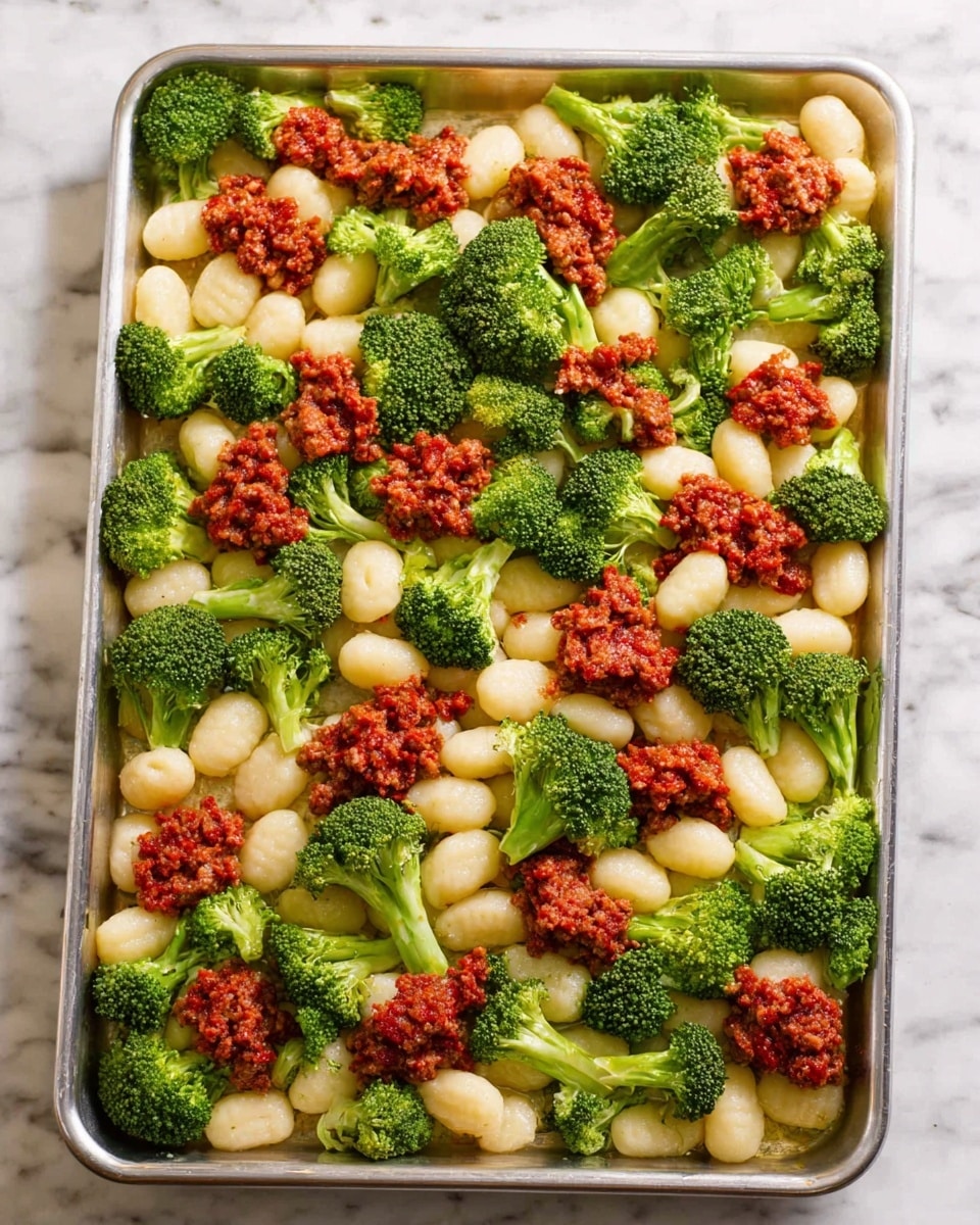 A large metal tray filled with three main layers spread evenly across its surface: white gnocchi pieces forming a soft, round base layer, bright green broccoli florets with textured tops and stalks placed in between, and small evenly spaced dollops of chunky red meat mixture on top. The tray rests on a white marbled texture. photo taken with an iphone --ar 4:5 --v 7