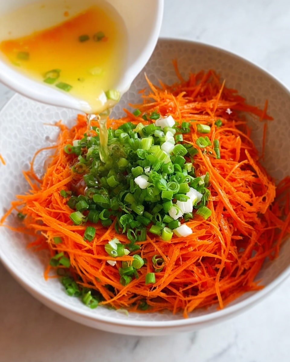 The image shows a white bowl on a white marbled surface filled with thin orange shredded carrot strands that create the main bottom layer. On top of the carrots, there are small green chopped scallions scattered around the center in a small heap that adds vibrant green color. In the next frame, a liquid, likely oil, is being poured from a pan over the vegetables, causing a slight shine and wet look on the carrots and scallions. The bowl is shown from a close angle with clear focus on the texture of the shredded carrots and fresh green scallions. Photo taken with an iphone --ar 4:5 --v 7