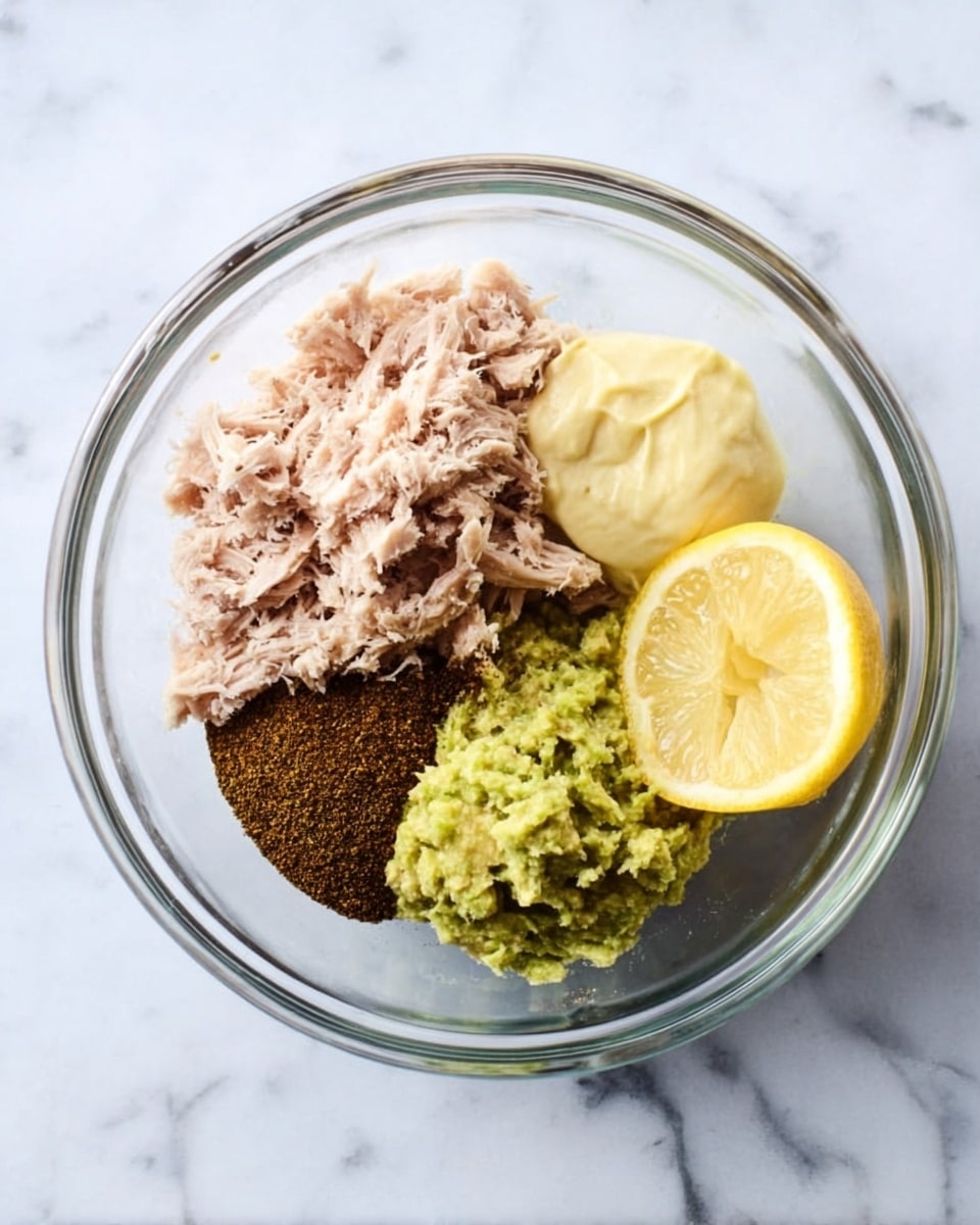 A clear glass bowl is placed on a white marbled surface, holding five distinct layers arranged side by side. Starting on the left, there is a light pink shredded meat layer with a soft texture. Next to it is a pile of finely ground dark brown spice. Above the spice, a thick pale yellow creamy layer sits in a dollop shape. To the right of the spice is a chunky green layer with a mashed texture. Finally, on the far right, there is a round slice of lemon with a yellow rind and pale yellow inside. Photo taken with an iphone --ar 4:5 --v 7
