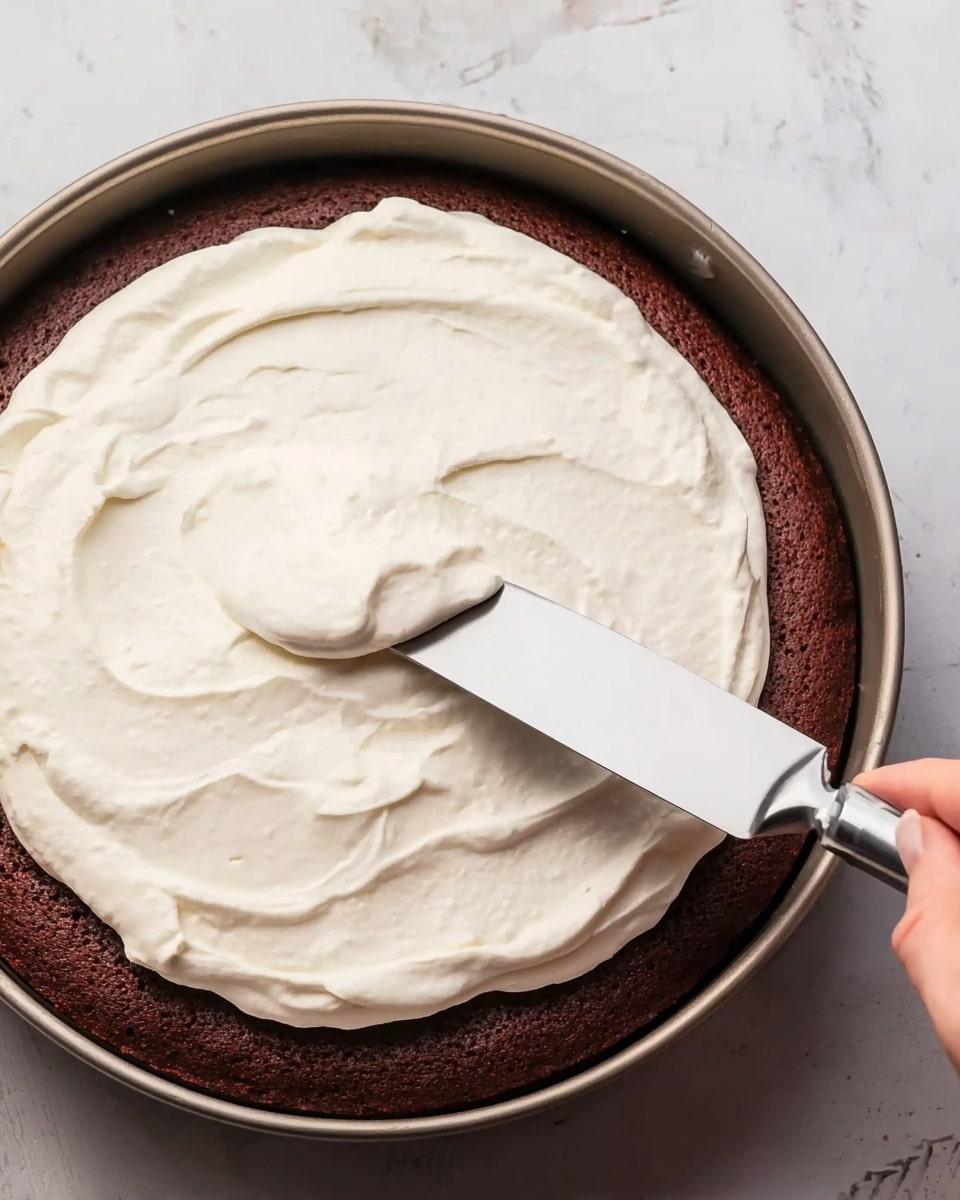 The image shows a baked chocolate cake in a round pan. The cake layer is dark brown and looks soft, taking up the bottom space of the pan. On top of this cake is a thick layer of white cream being spread evenly with a metal spatula. The spatula is held by a woman's hand from the right side. The background is a white marbled texture. photo taken with an iphone --ar 4:5 --v 7