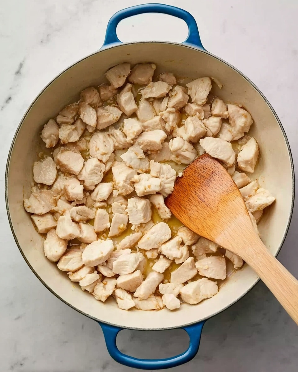 A white pot with blue handles is filled with small pieces of light beige chicken cooking inside. The chicken pieces are spread out evenly, showing a slight shine from cooking oils. A wooden spatula with a smooth texture is resting in the pot, partially under some chicken pieces, angled towards the bottom right. The pot is placed on a white marbled surface. photo taken with an iphone --ar 4:5 --v 7