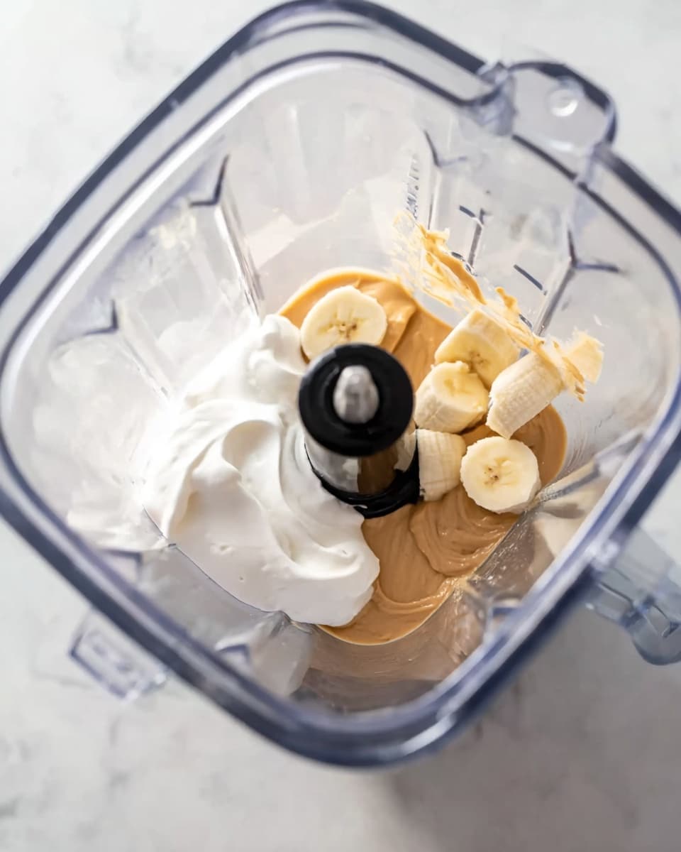 Inside a clear blender container on a white marbled surface, three main layers sit ready to blend: at the bottom is a light beige peanut butter layer with a smooth and creamy texture, beside it lies two pieces of sliced banana with pale yellow color and soft, slightly textured surface, and atop these is a dollop of white creamy yogurt with a fluffy texture. The blender's central blade and rod stand upright in the middle, casting slight shadows on the ingredients. photo taken with an iphone --ar 4:5 --v 7
