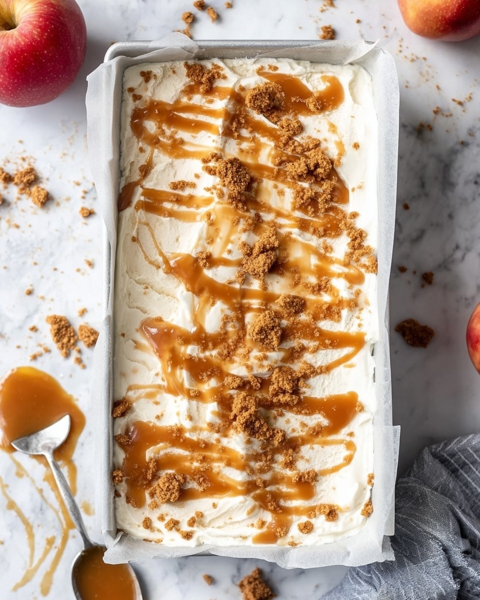 The image shows a rectangular ice cream dessert in a white tray lined with white parchment paper. The base layer is creamy white ice cream with a soft, swirled texture. On top, there are uneven drizzles of light brown caramel sauce creating shiny, glossy streaks across the surface. Crumbled brown cookie pieces are scattered over the caramel, adding a crumbly texture on top. The tray rests on a white marbled surface with some spilled caramel and cookie crumbs nearby. Two red apples are visible in the blurred background on the left side. A spoon with caramel sauce drips from it is placed on the lower left corner. photo taken with an iphone --ar 4:5 --v 7