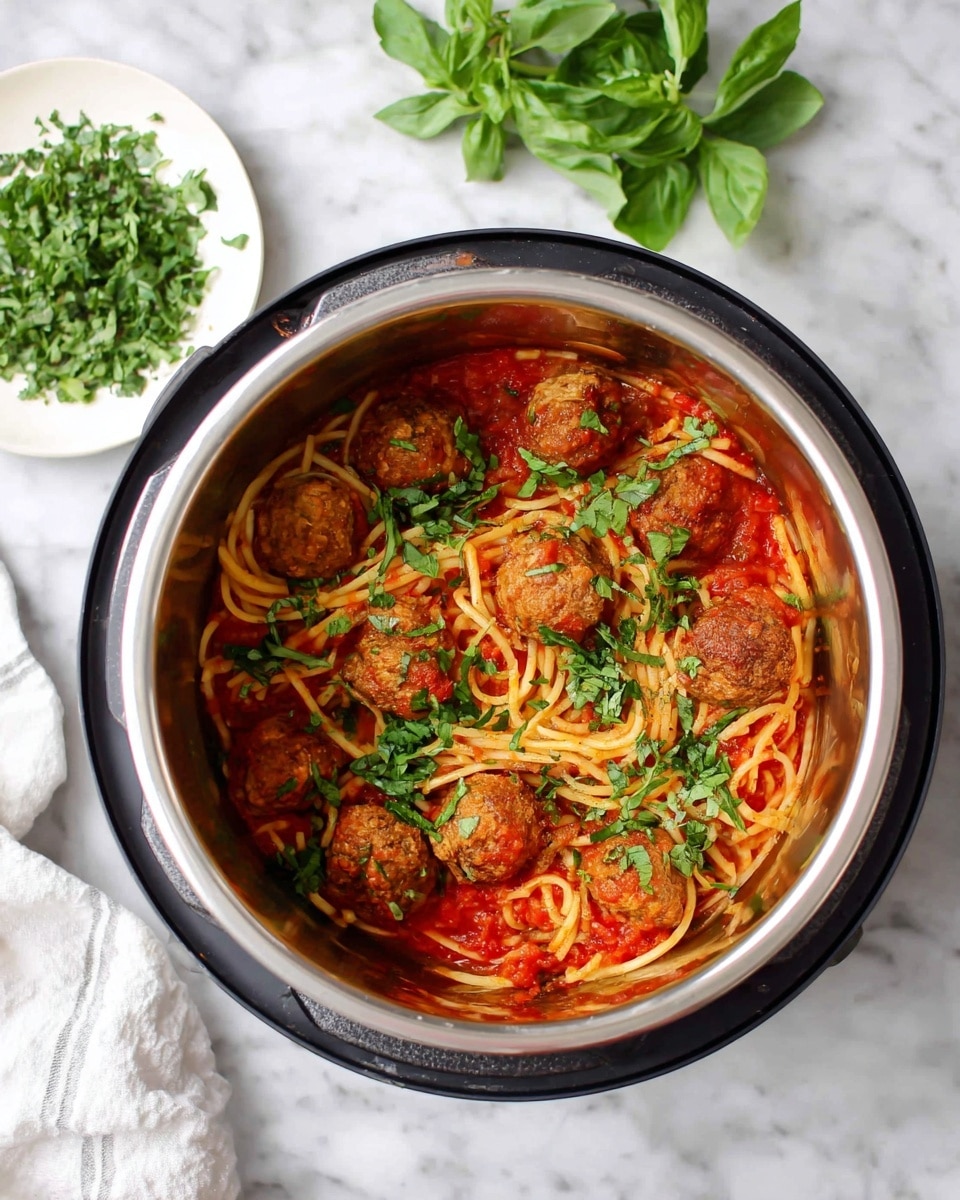 Inside a metal pot, there is a dish with two main layers: the bottom layer is spaghetti noodles mixed with red tomato sauce, showing smooth and slightly shiny texture, while on top there are several round, brown meatballs scattered evenly. Bright green chopped herbs are sprinkled over the meatballs and noodles, adding fresh color contrast. The pot sits on a white marbled surface along with a white plate holding more chopped green herbs and fresh basil leaves nearby. Photo taken with an iphone --ar 4:5 --v 7