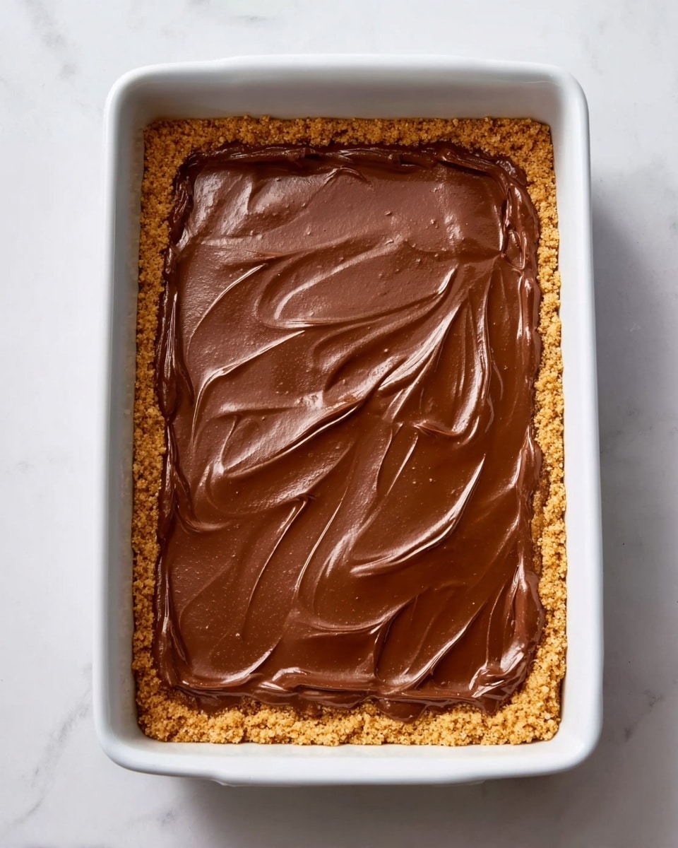 A rectangular white baking dish holds a dessert with two visible layers: the bottom layer is a rough, golden-brown crust that lines the dish entirely, and the top layer is a smooth, shiny chocolate spread that covers the crust evenly, with gentle swirls on the surface. The dish is placed on a white marbled textured surface. Photo taken with an iphone --ar 4:5 --v 7