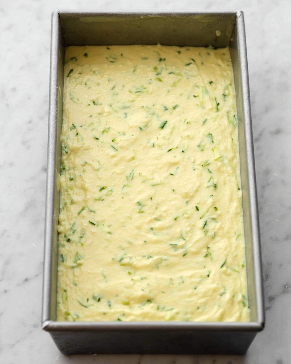 A close-up view of a rectangular metal baking pan filled with a light yellow batter that has small green specks scattered throughout, indicating finely shredded vegetables mixed in. The batter is spread evenly, covering the entire bottom of the pan with a smooth but slightly textured surface. The pan is placed on a white marbled textured surface. Photo taken with an iphone --ar 4:5 --v 7
